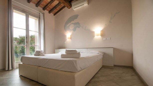 A bedroom with a large bed, a window with curtains, a wall-mounted air conditioner, and a wall with a decorative light fixture. - Limonaia , one-bedroom terraced house (Cecina)