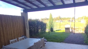 Eine Terrasse im Freien mit Tisch und Stühlen, einem überdachten Bereich und Blick auf eine Wiese und Bäume.
