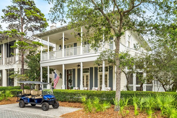 A two-story building with a porch, a flag, and a golf cart. - Lake District Luxury: 6-Bedroom Retreat with WaterColor Beach Club Access, Bikes, and Electric Cart! (Santa Rosa Beach)