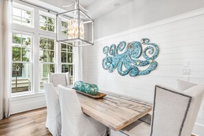 A dining area with a wooden table, white chairs, and a decorative wall piece. - Lake District Luxury: 6-Bedroom Retreat with WaterColor Beach Club Access, Bikes, and Electric Cart! (Santa Rosa Beach)
