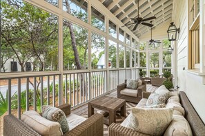 A covered porch with wicker furniture, a wooden table, and a view of the surrounding trees and street.