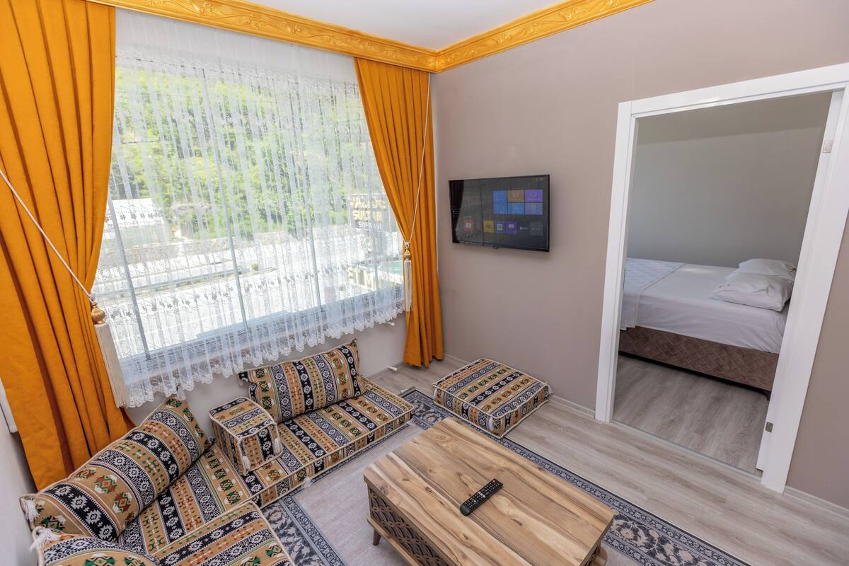 Une pièce comprenant un ensemble de canapés, une table basse en bois, un téléviseur fixé au mur et un lit dans la pièce adjacente.