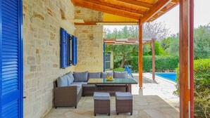 Un'area relax esterna coperta con parete in pietra, persiane blu e piscina sullo sfondo.