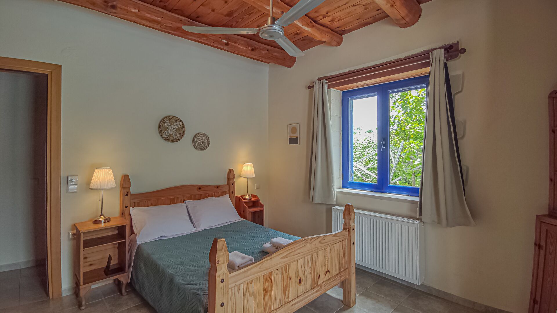 A bedroom with a wooden bed, two bedside tables with lamps, a ceiling fan, a window with curtains, and a radiator.