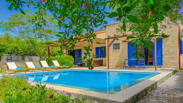 Una piscina con lettini, un edificio in pietra con persiane blu e un pergolato con piante.