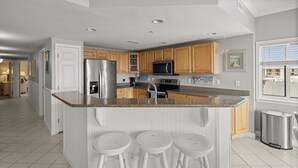 A modern kitchen with wooden cabinets, stainless steel appliances, and a granite countertop. There are three white stools at the island. - Avalon 507-Oceanside 140th St, Free WiFi, Pool, Elev, W/D, AC (Ocean City)