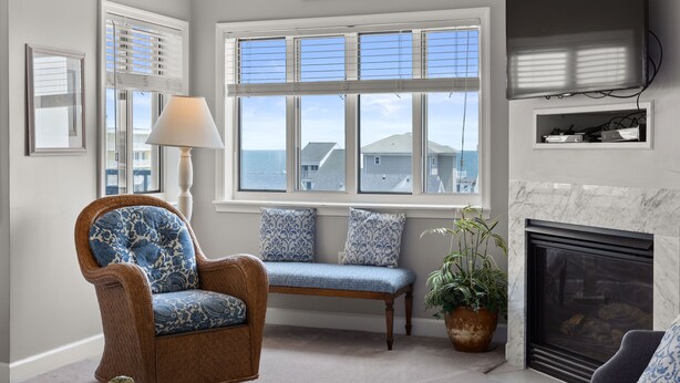 A living room with a wicker chair, a bench, a fireplace, and a large window with blinds. - Avalon 507-Oceanside 140th St, Free WiFi, Pool, Elev, W/D, AC (Ocean City)