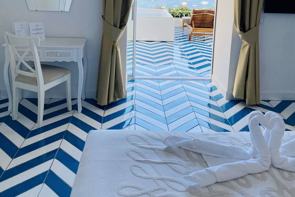 Ein Hotelzimmer mit Bett, Schreibtisch und Stuhl. Im Hintergrund ist Meer und eine Berglandschaft zu sehen.