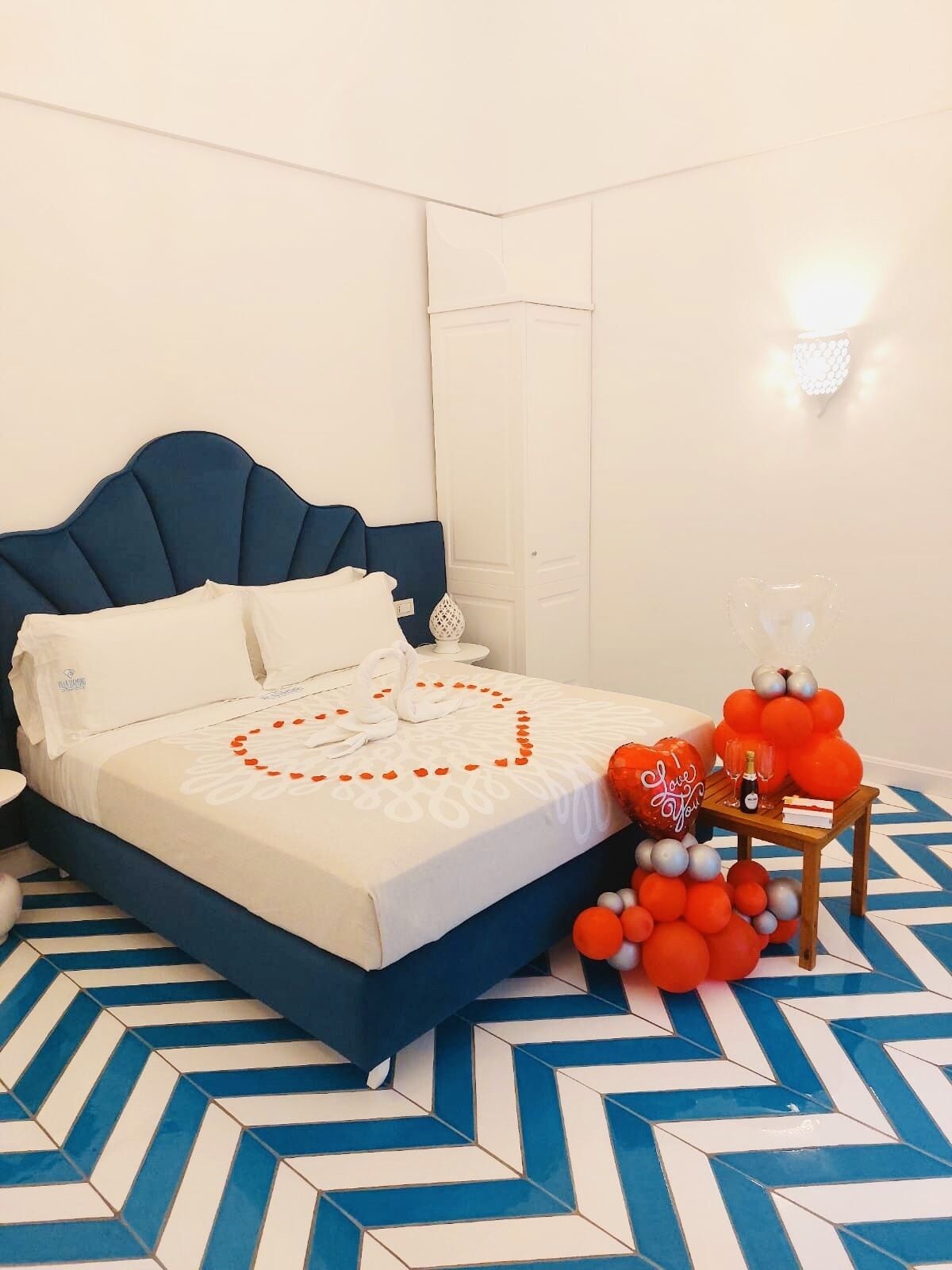 A neatly made bed with a heart-shaped decoration, a small table with balloons and a bottle, and a blue and white patterned floor.