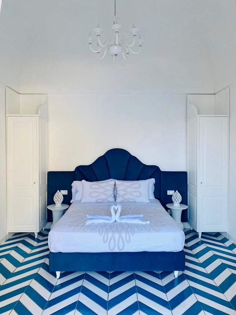 A bed with a dark headboard, white bedding, and a heart-shaped towel arrangement. There are two bedside tables with decorative items. The floor has a blue and white chevron pattern.