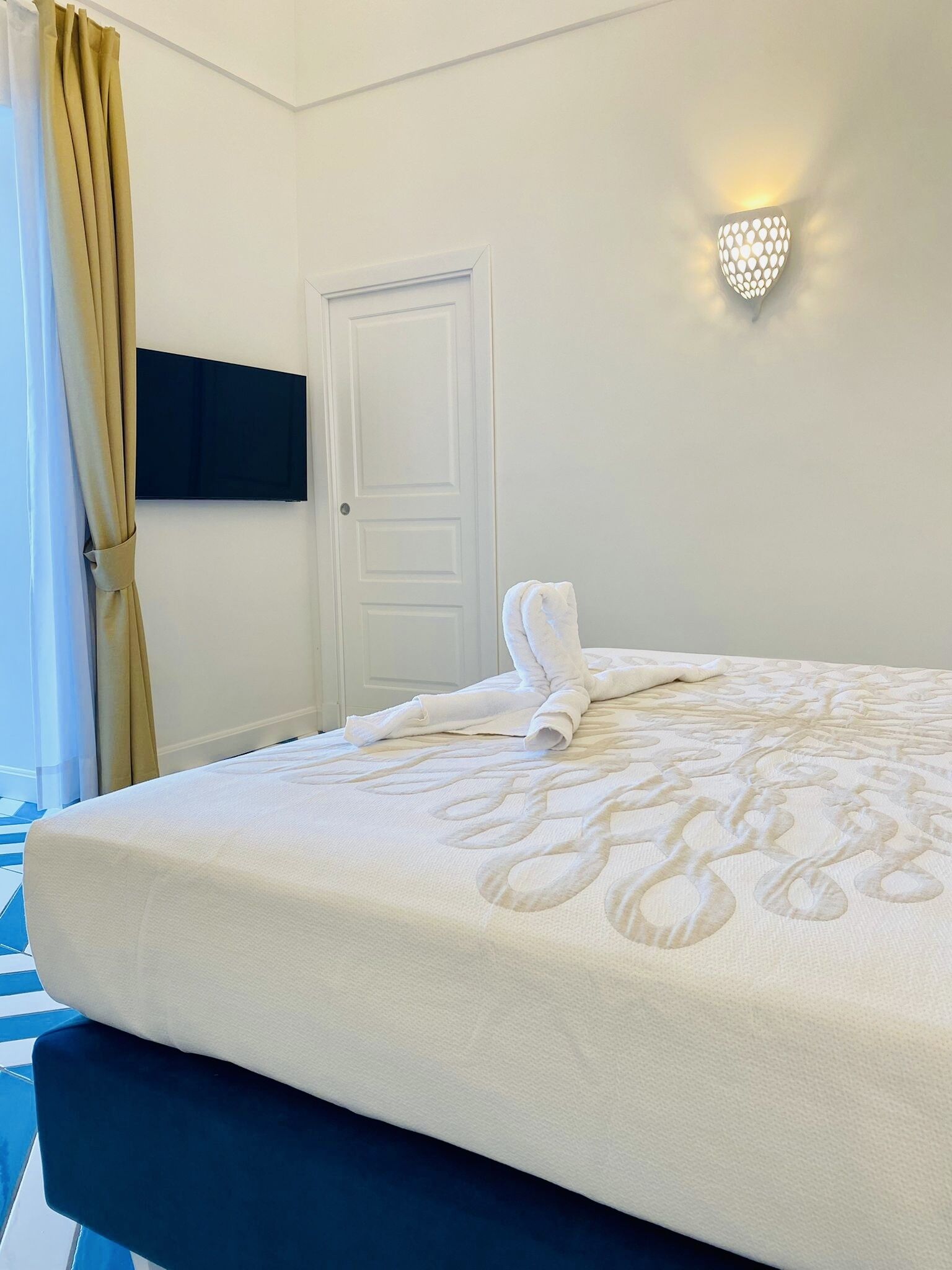 A hotel room with a bed, a flat-screen TV mounted on the wall, a door, and a wall-mounted light fixture.