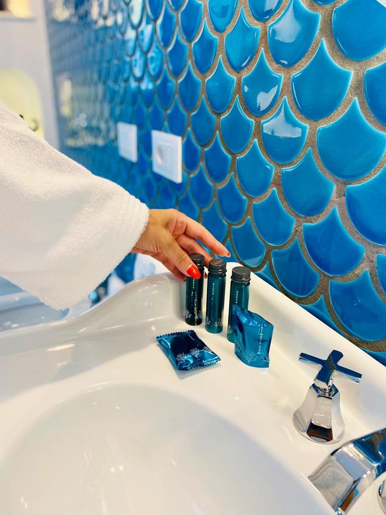 A hand placing a blue bottle into a sink with a white countertop and a blue tiled wall in the background.
