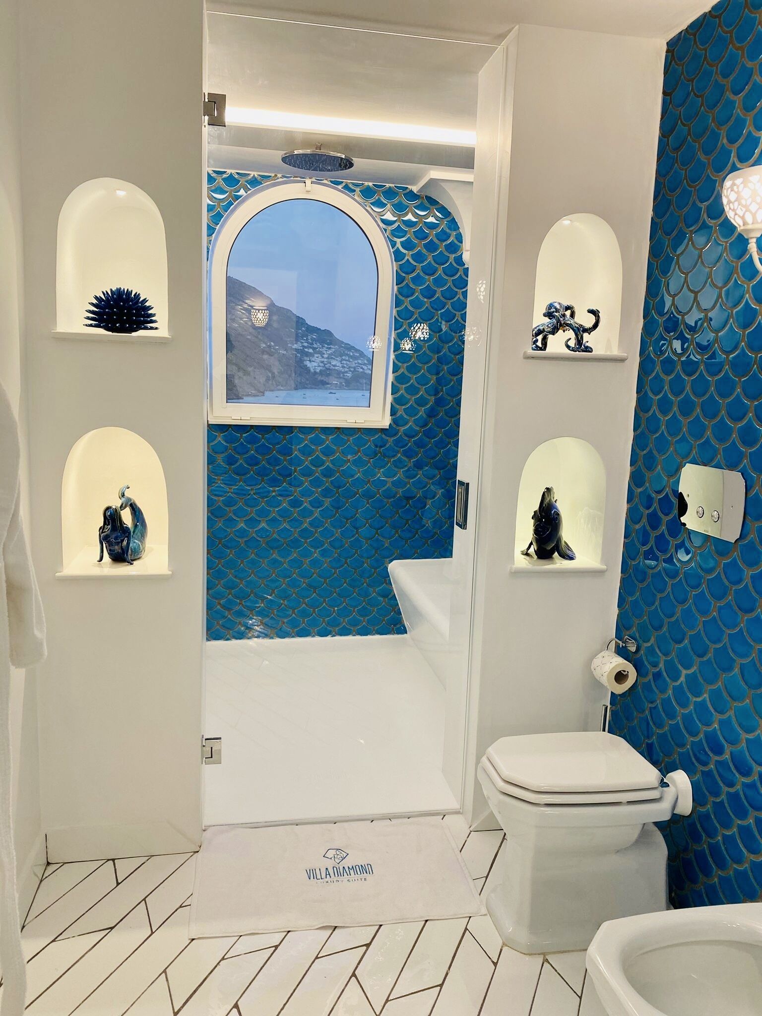 Une salle de bain avec un mur recouvert de carreaux bleus, une fenêtre donnant sur les montagnes, des toilettes et un lavabo.