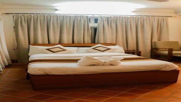 A neatly made bed with pillows and a blanket, a chair, a small table with a lamp, and a window with curtains. - MeroStay 124 (Kathmandu)