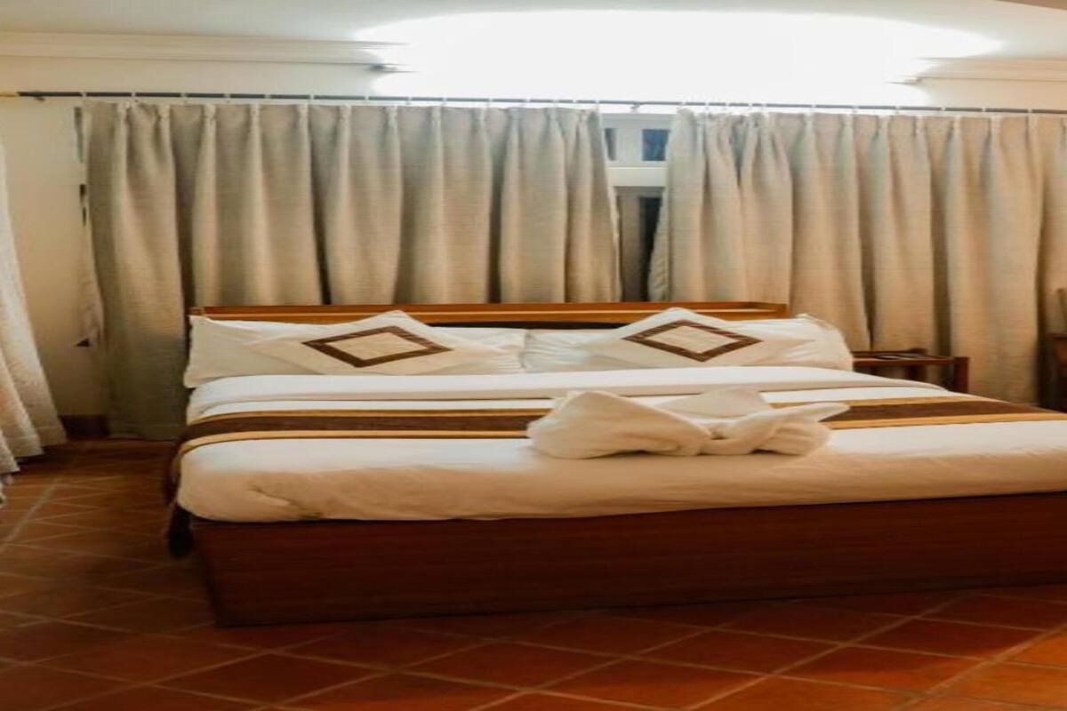 A neatly made bed with pillows and a blanket, a chair, a small table with a lamp, and a window with curtains.