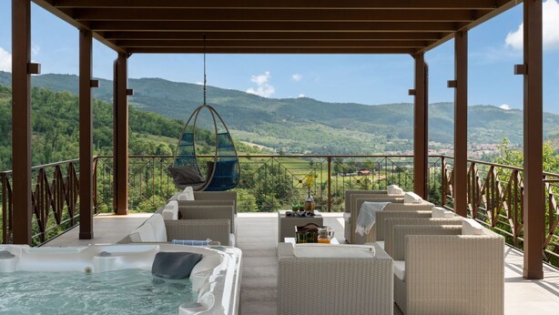 Un espacio al aire libre techado con mobiliario de mimbre, jacuzzi y una silla colgante con vistas a un paisaje montañoso.