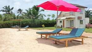 A beachside resort with wooden lounge chairs and a red umbrella, surrounded by palm trees and a white building with a balcony. - La Villa Aphro Benin (Ouidah)