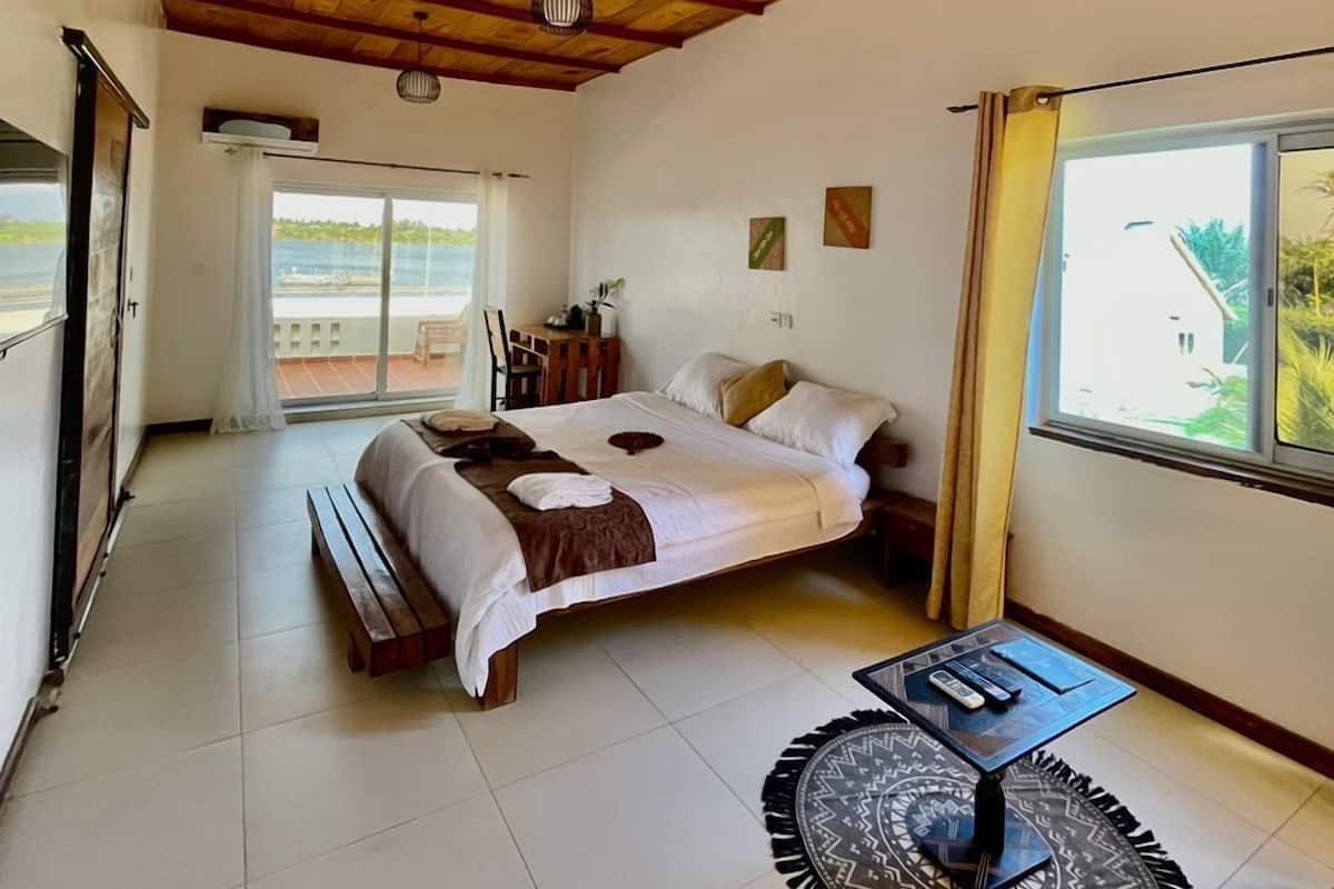 A spacious bedroom with a large bed, a round coffee table, and a view of the outdoors through a large window.