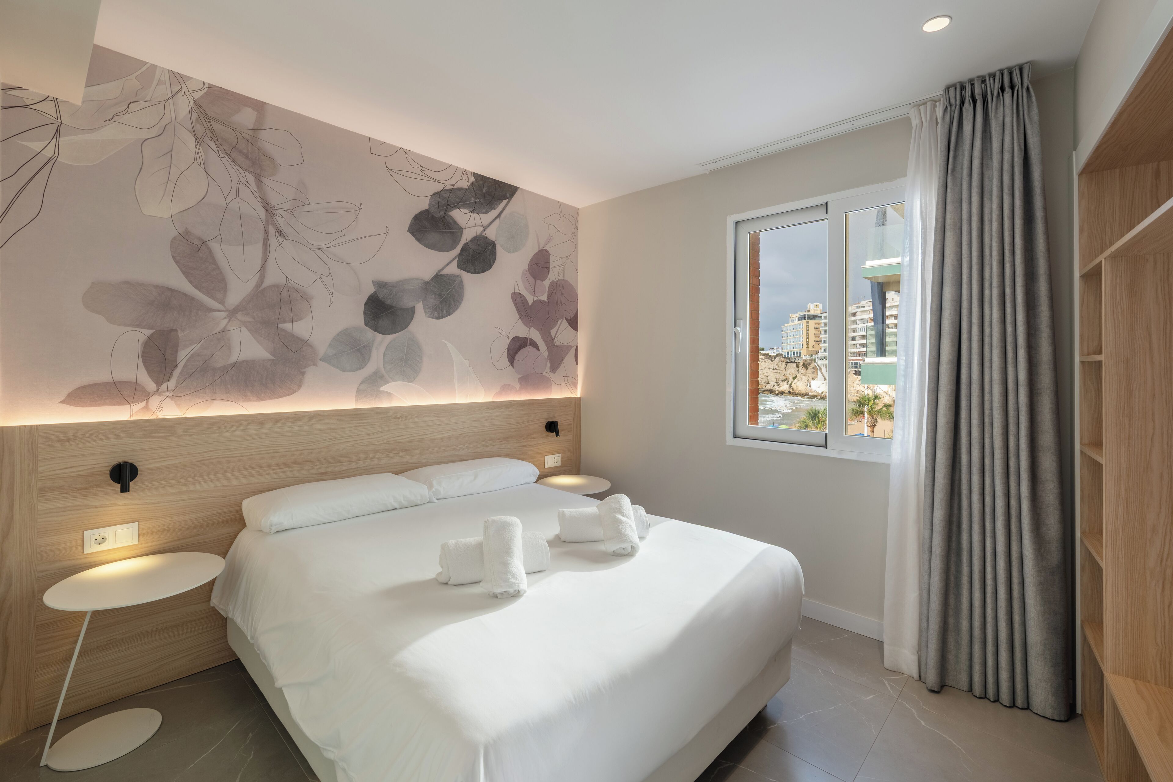 Habitación de hotel moderna con una cama grande, mesitas de noche, un mural en la pared, una ventana con cortinas y vistas a edificios.