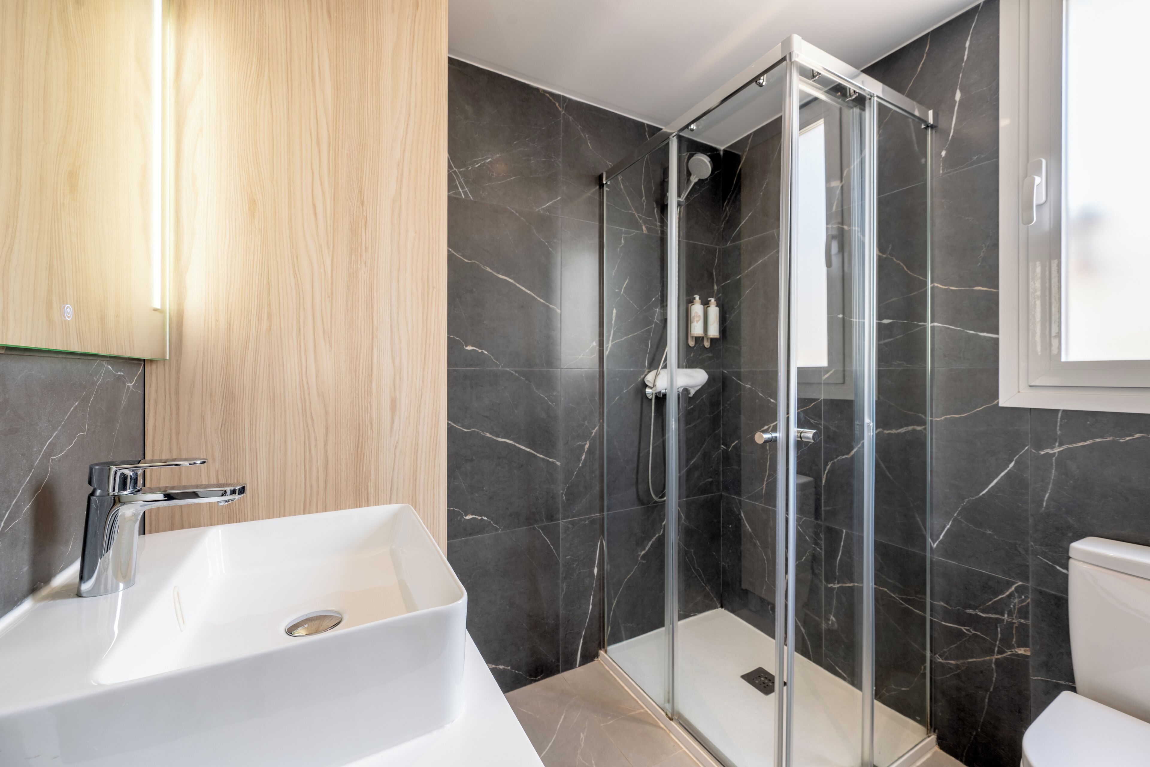 Une salle de bain moderne avec une douche à parois de verre, un lavabo blanc et des murs en marbre foncé.