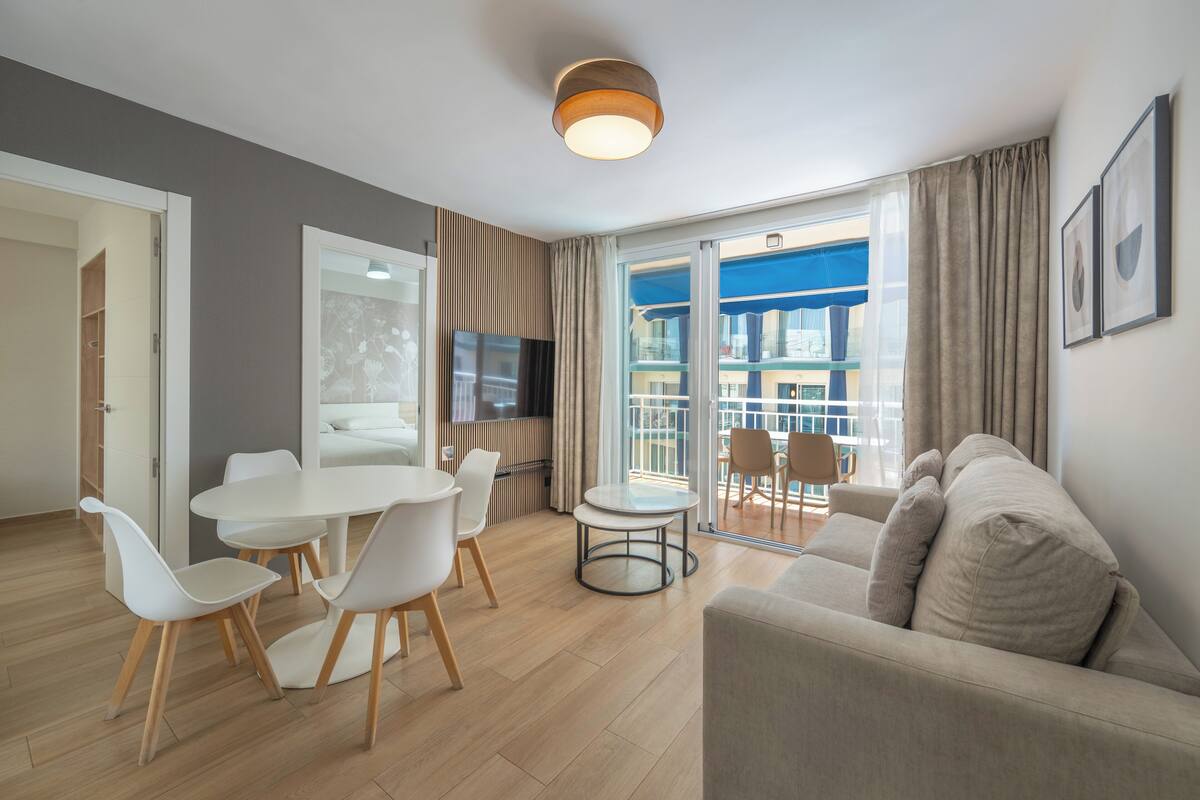 A modern living room with a sofa, dining table, and chairs, a TV, and a balcony with a view.