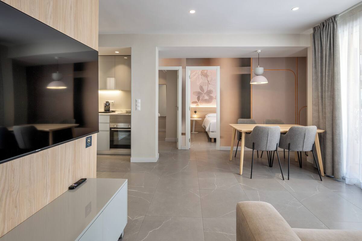 A modern hotel room with a flat-screen TV, a dining area with a table and chairs, and a separate bedroom visible through an open door.