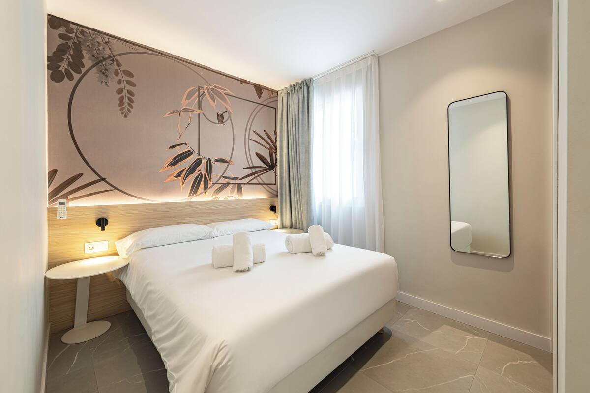 A hotel room with a bed, a bedside table, a wall-mounted mirror, and a decorative wall panel.