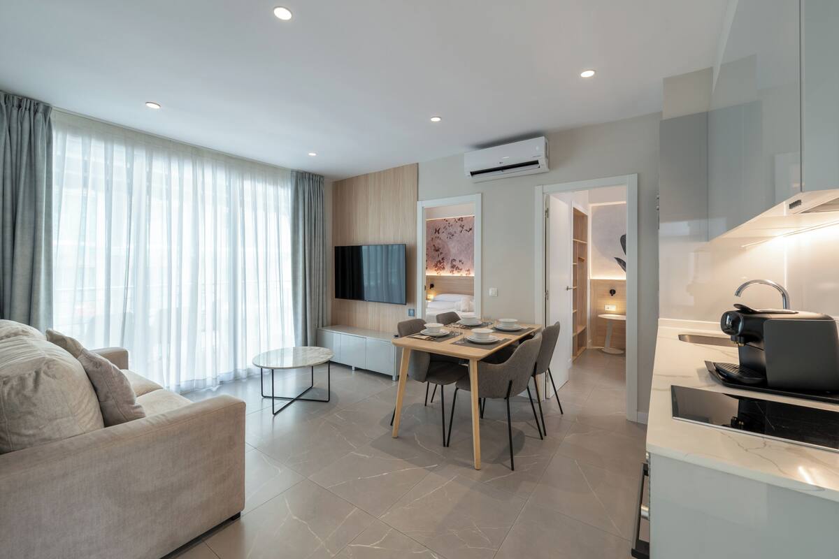 A modern living room with a sofa, a dining table with chairs, a TV mounted on the wall, and an air conditioning unit.