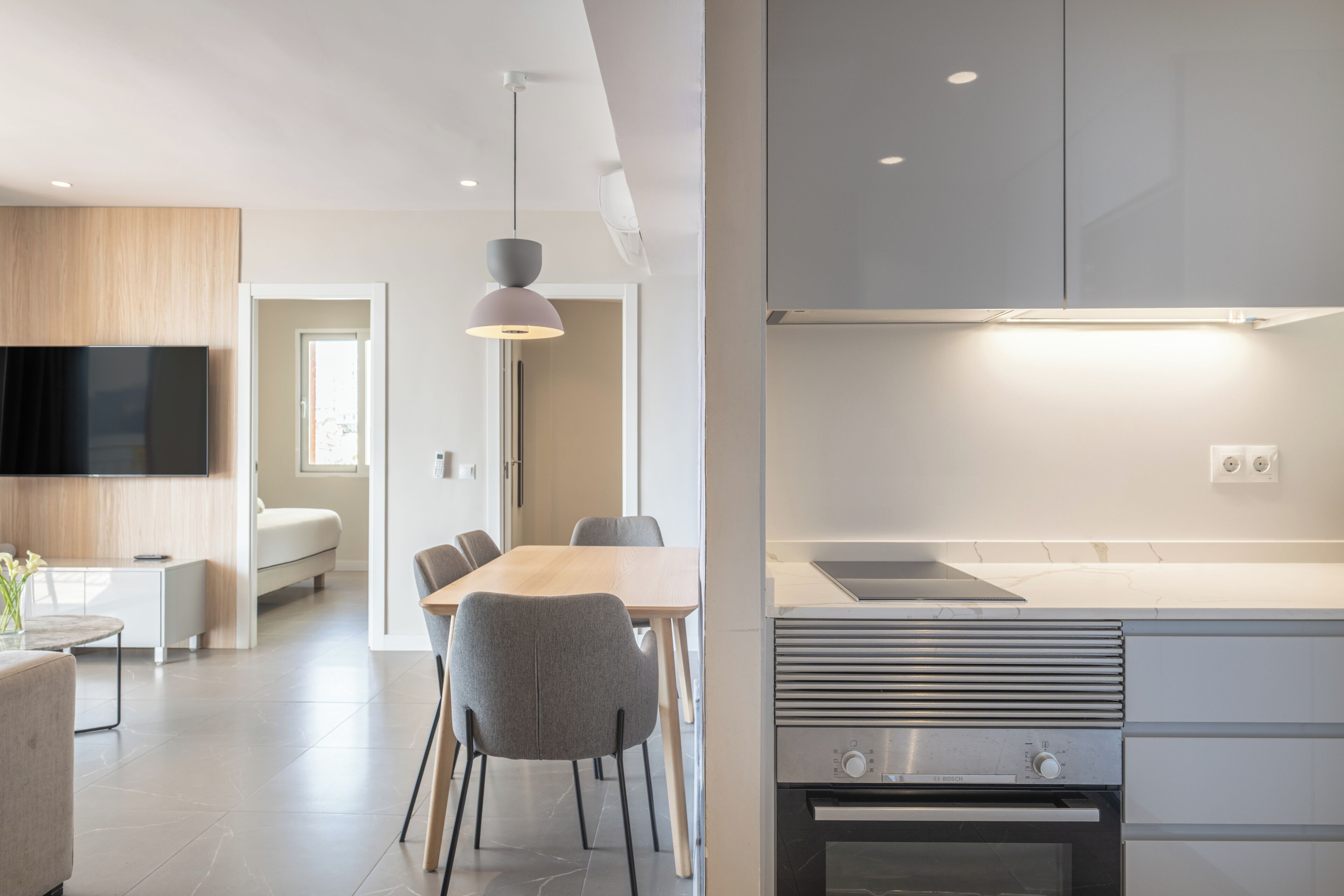 A modern kitchen with a Bosch oven, a dining area with a wooden table and grey chairs, and a flat-screen TV mounted on the wall.