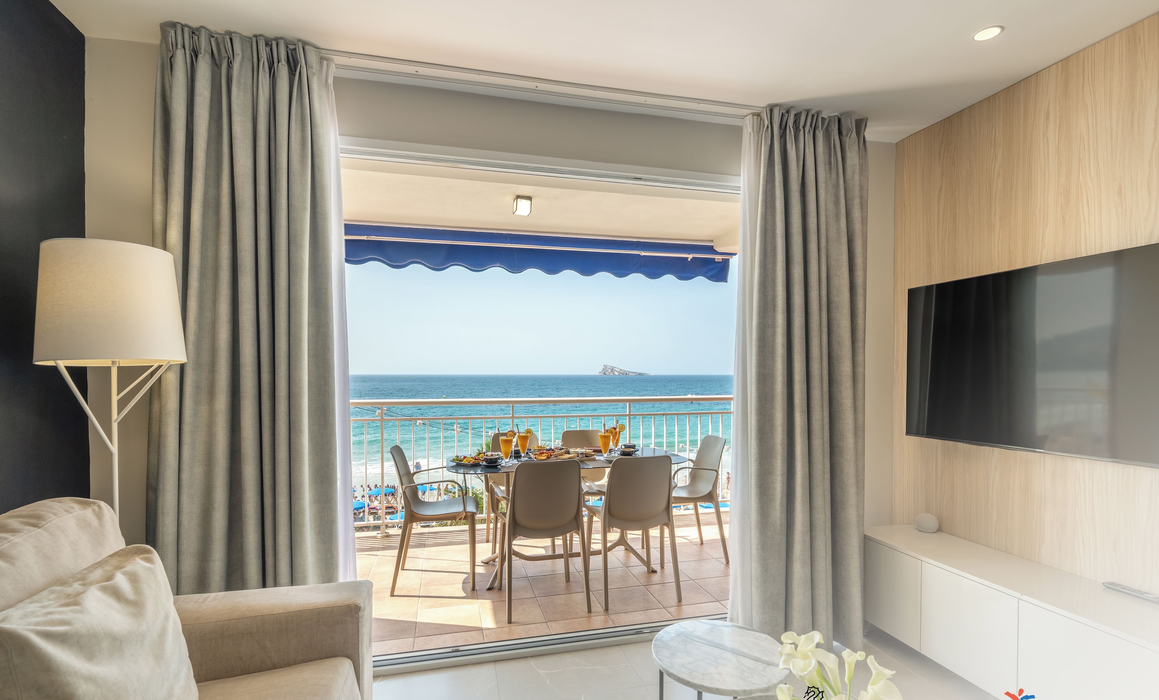 Habitación de hotel moderna con balcón con vistas al mar, zona de comedor con mesa para dos personas y televisor de pantalla plana montado en la pared.