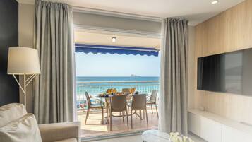 Habitación de hotel moderna con balcón con vistas al mar, zona de comedor con mesa para dos personas y televisor de pantalla plana montado en la pared.