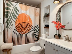 Ein Badezimmer mit Duschvorhang mit Blättermotiv, Toilette, Waschbecken mit Spiegel und einem kleinen Hocker mit Handtuch.