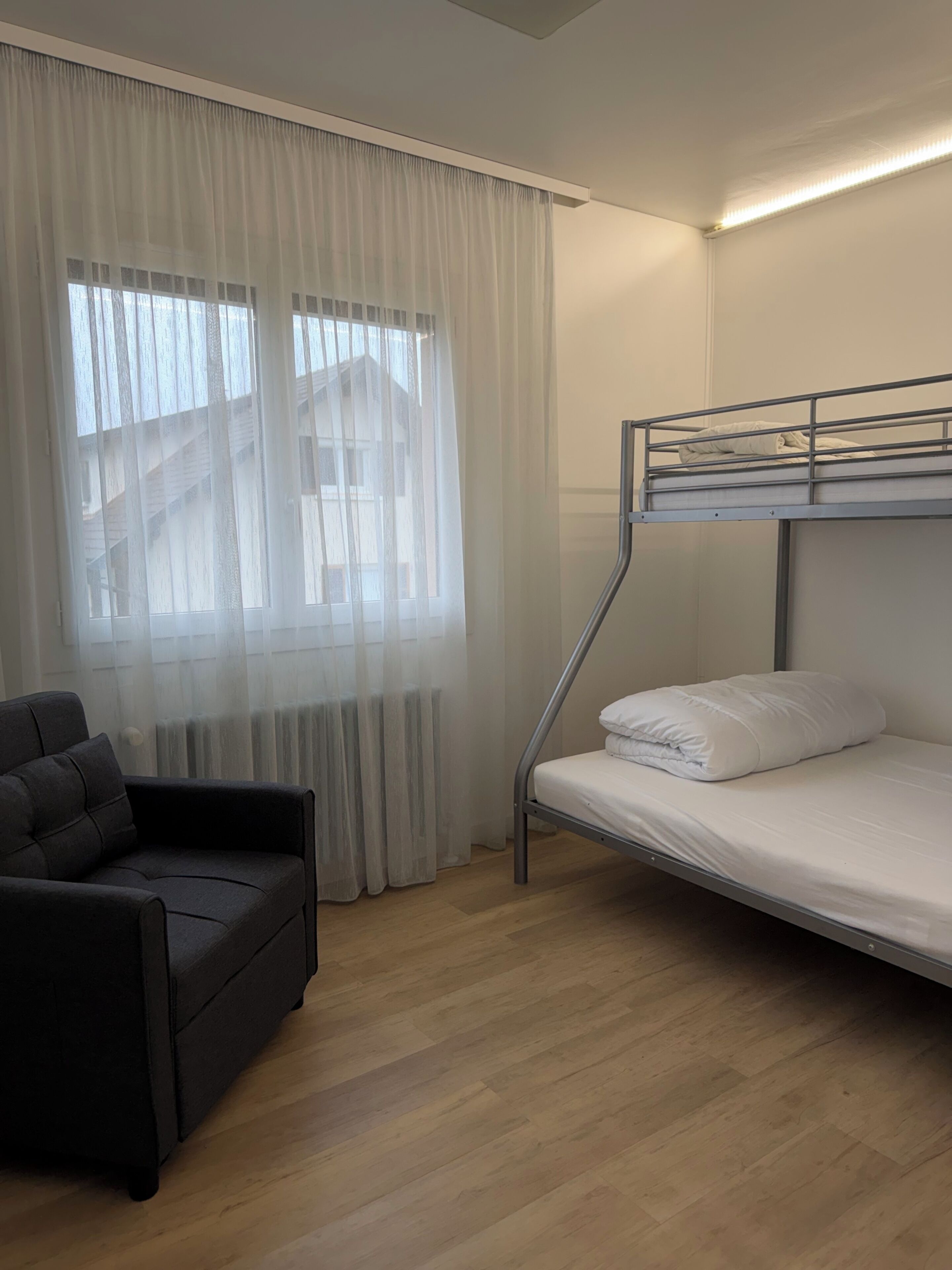 Una habitación con literas, un sofá, un ventanal con cortinas traslúcidas y un radiador.