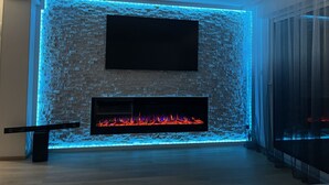Ein modernes Wohnzimmer mit Steinwand, einem flachbildfernseher über einem Kamin mit blauer Beleuchtung und einem Glasabtrennung auf der rechten Seite.