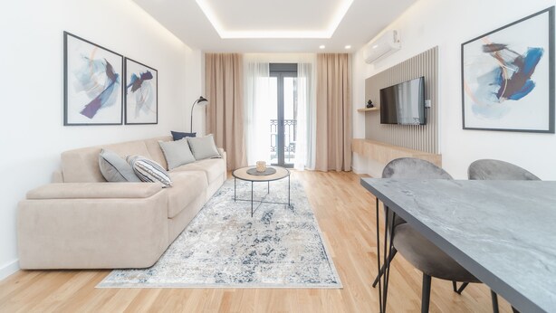 A modern living room with a sofa, coffee table, and dining table. There is a TV mounted on the wall and a balcony visible through the window. - Salus Hill Condo (Budva)