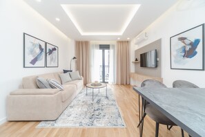 Un salon moderne avec un canapé, une table basse et une table à manger. Un téléviseur est fixé au mur et un balcon est visible à travers la fenêtre.