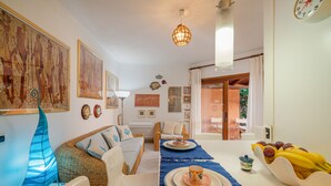 A cozy living room with a sofa, chairs, a dining table with plates and a fruit bowl, and artwork on the walls. - Casa Flavia by Wonderful Italy (Golfo Aranci)