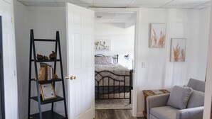 A bedroom with a bed, a gray armchair, a black shelf with books, and a door leading to another room. - Quiet farm stay, hike, mushroom hunt, see the animals, relax, so much more! (Edmonton)