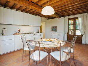 Una cocina con armarios blancos, una mesa redonda para comer con cuatro sillas, una lámpara colgante y una ventana con cortinas.