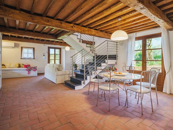 Ein geräumiger Wohnbereich mit Kamin, einem Esstisch mit Stühlen, einer Treppe und einem großen Fenster mit Vorhängen.