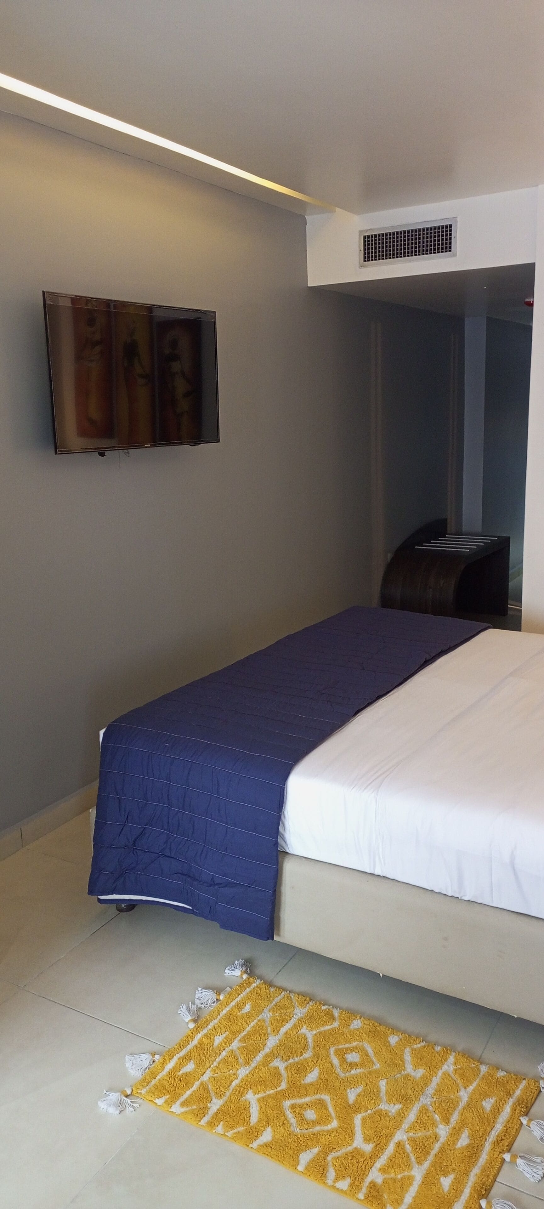 Camera d'albergo con un letto, una TV a parete, una bocchetta di ventilazione e un tappeto giallo fantasia.