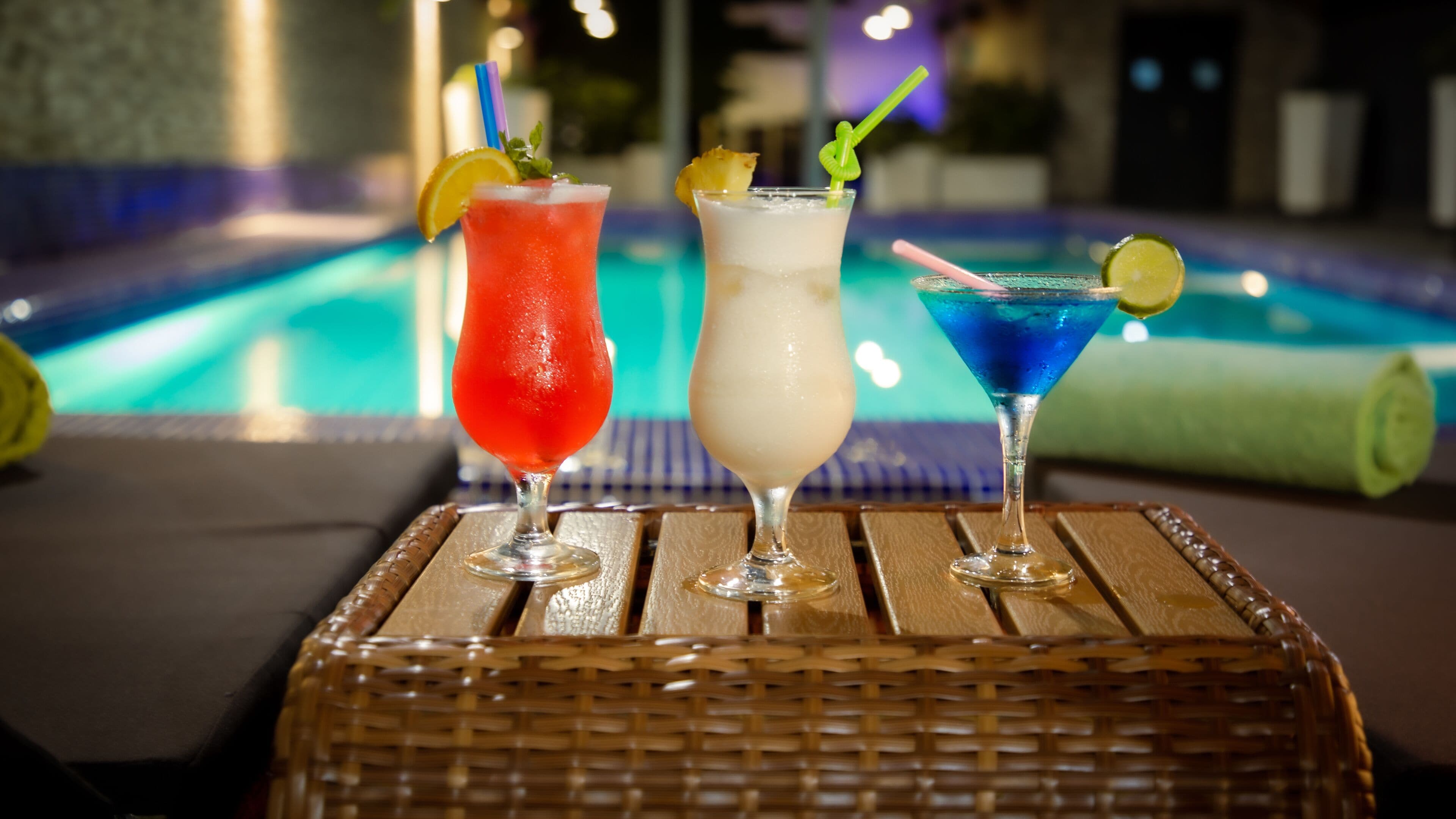 Three cocktails on a wooden table by a poolside.