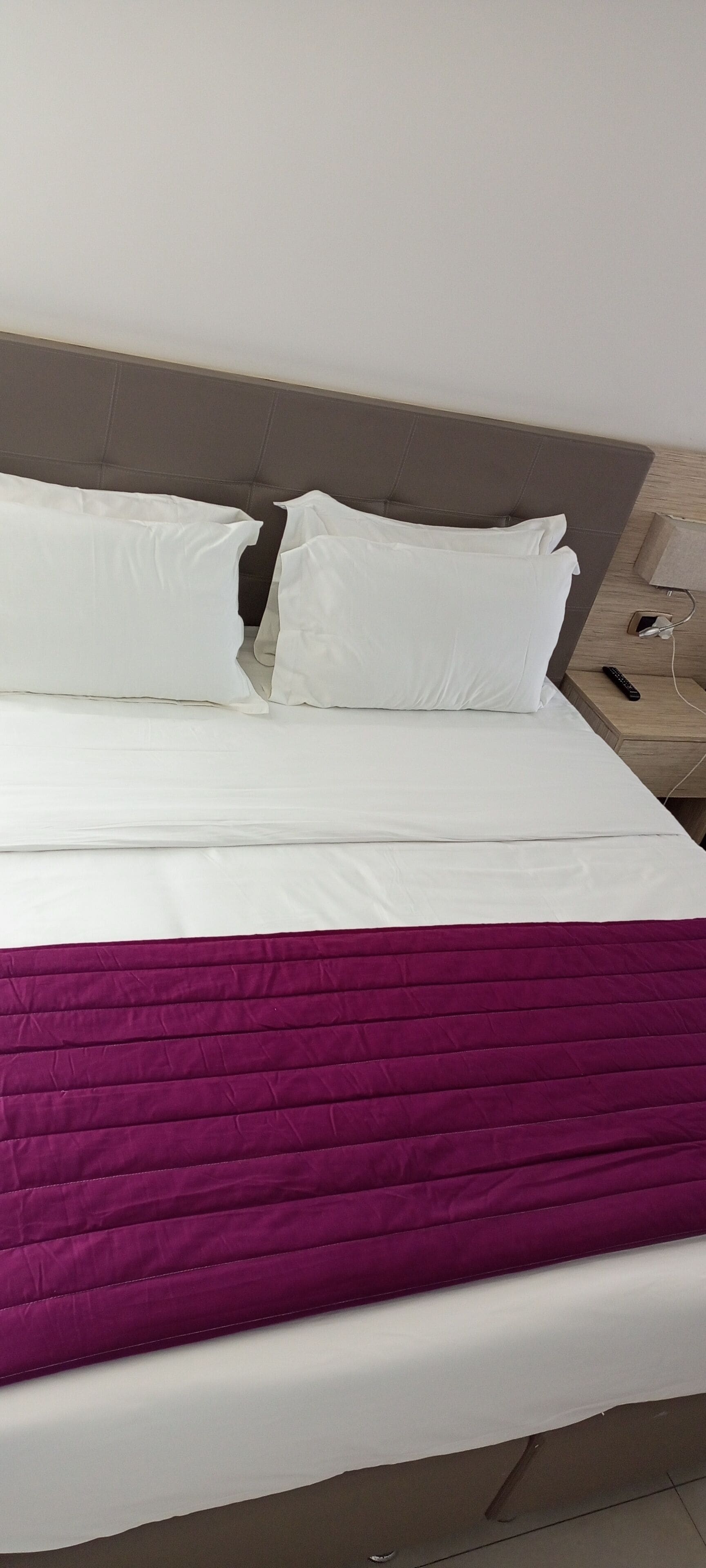Una cama con ropa de cama blanca y morada, un cabecero gris y una mesita de noche con una lámpara y un control remoto.
