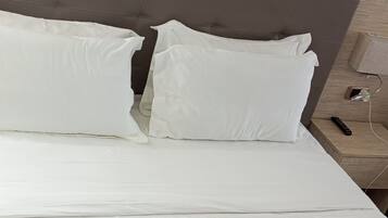 Una cama con ropa de cama blanca y morada, un cabecero gris y una mesita de noche con una lámpara y un control remoto.