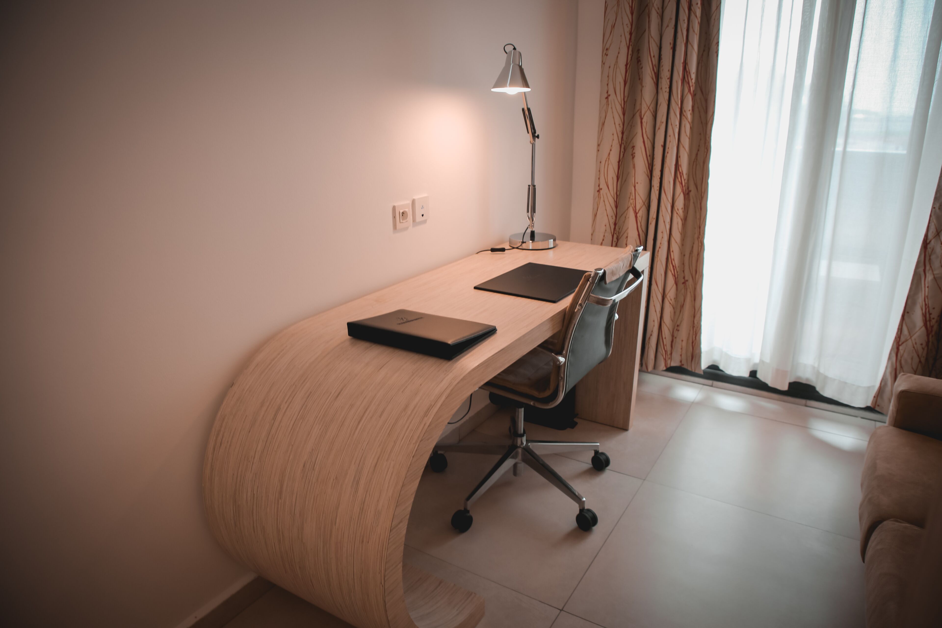 A modern office space with a curved desk, a chair, a desk lamp, and a window with curtains.