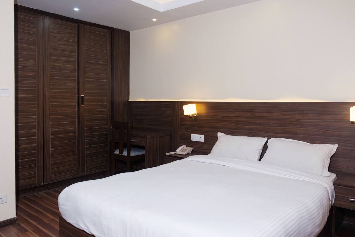 A hotel room with a large bed, wooden wardrobe, bedside table with a lamp, and a small desk with a chair.