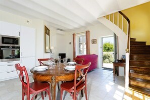A dining area with a wooden table and red chairs, a kitchen with white cabinets and stainless steel appliances, and a staircase leading to a balcony with a view of greenery.