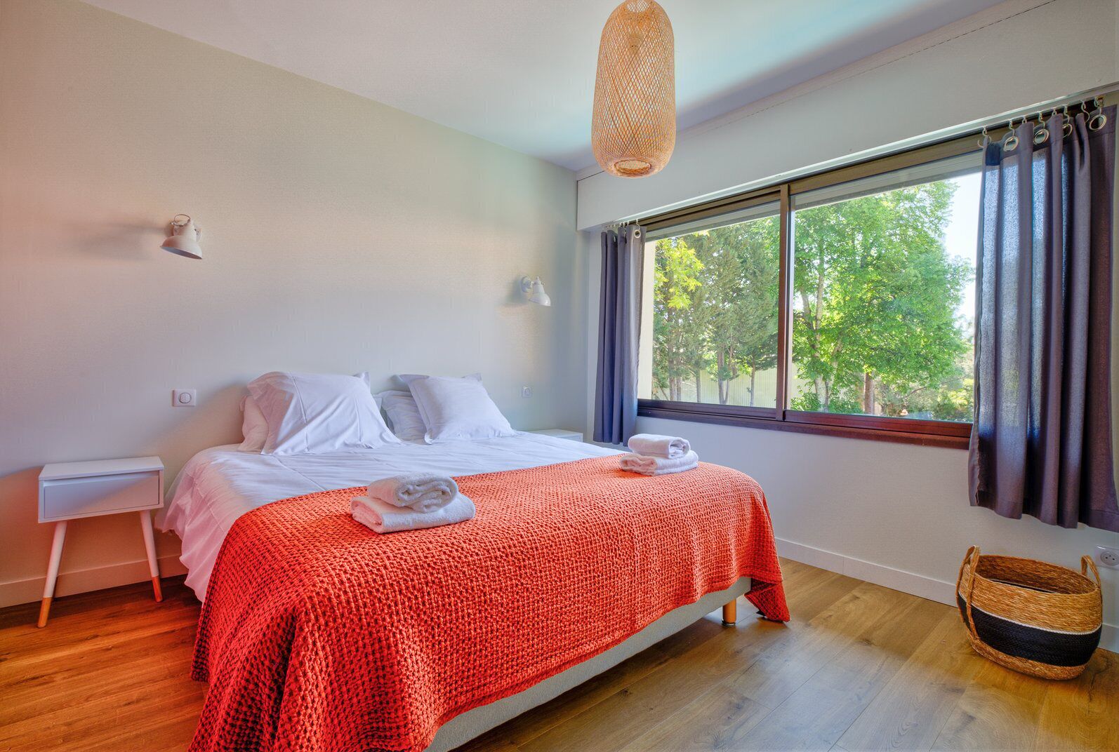 A bedroom with a bed, a nightstand, a window with curtains, and a wicker basket.