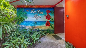 En väggmålning med ett tropiskt strandmotiv och hibiskusblommor, en röd dörr och frodig grönska.