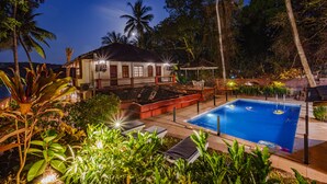 Un área de piscina con sillones de descanso, una casa con balcón y vegetación tropical.